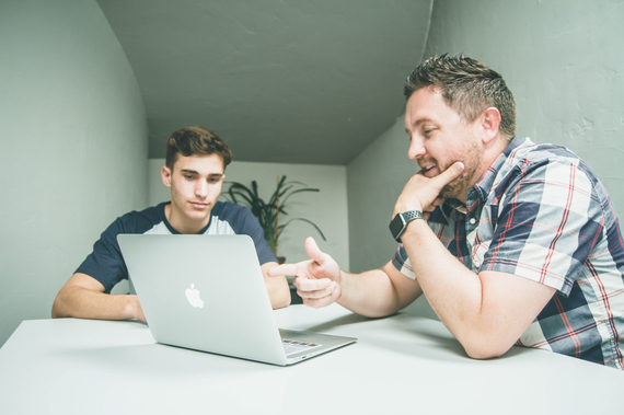 Zwei Männer sitzen an einem Tisch und schauen auf einen Laptop.