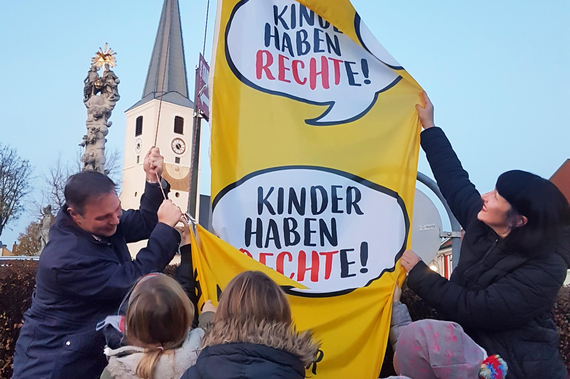 Bürgermeister Andreas Babler hisst gemeinsam mit Karin Blum und Kindern die Kinderrechte-Fahne.